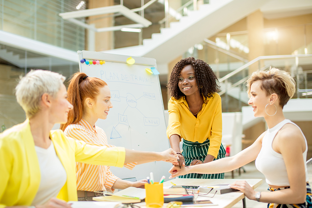 Cuatro mujeres se unen con las manos en un gesto de equipo, sonriendo en una oficina.